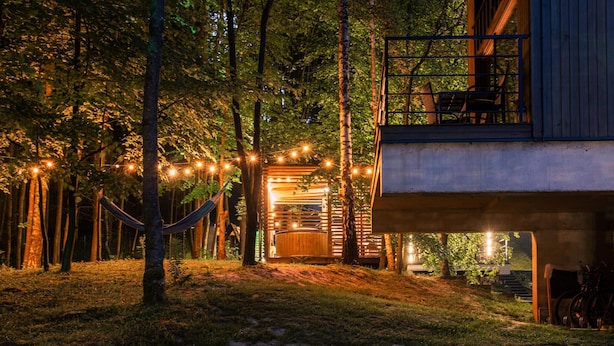 A modern building with a balcony overlooking a forested area. There is a hammock strung between trees and string lights illuminating the path.
