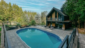 A modern house with a swimming pool, surrounded by a wooden deck and a forest.