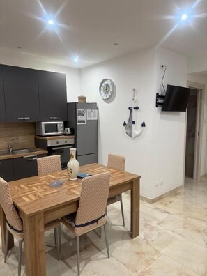 A modern kitchen with dark cabinets, a microwave, and a refrigerator. There is a dining table with chairs and a vase on it. A flat-screen TV is mounted on the wall. - Aphrodite's apartment at Aspra (BAGHERIA)