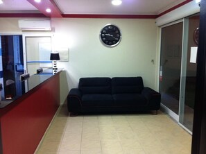 A living room with a black sofa, a wall clock, a small table with a lamp, and a sliding glass door. - Paraiso Dourado (Porto Santo)