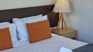 A neatly made bed with white and orange pillows, a wooden nightstand with a lamp, and folded white towels on the bed.