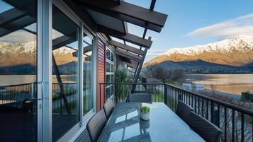 A balcony with a glass table and chairs, overlooking a mountainous landscape and a body of water.