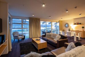 Una sala de estar moderna con un sofá, sillones, una mesa de centro y un televisor. Amplias ventanas ofrecen vistas al exterior.