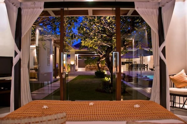 A bedroom with a large bed, a view of a swimming pool, and a tree outside the window. - Chandra Bali Villas (Seminyak)
