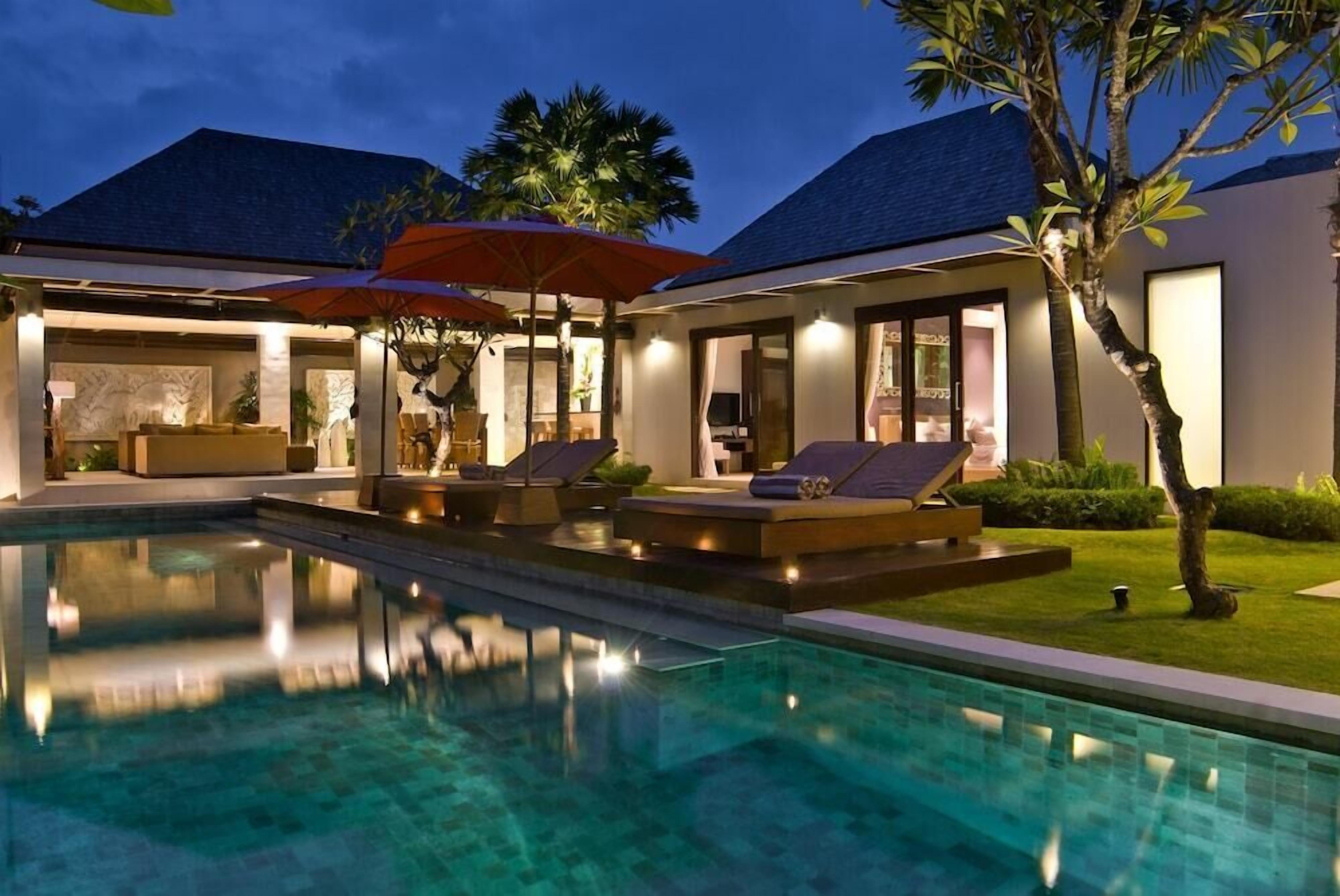 A swimming pool with lounge chairs and umbrellas, surrounded by a well-lit outdoor area with palm trees.