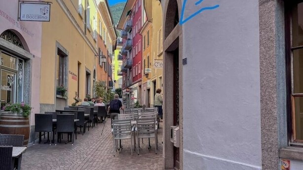 Una estrecha calle adoquinada flanqueada por edificios coloridos, mesas al aire libre de un restaurante y un cielo azul despejado.