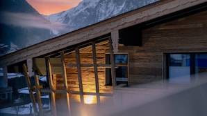 Un chalet en bois doté de grandes fenêtres et d’un balcon donnant sur une chaîne de montagnes enneigées.