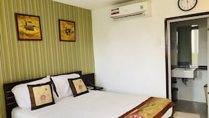 Habitación de hotel con cama, dos almohadas, una mesita de noche, equipo de aire acondicionado, un reloj y baño visible a través de una puerta abierta.