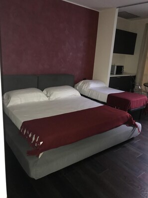 Et hotelværelse med to senge, mørkt gulv og et tekøkken i baggrunden.