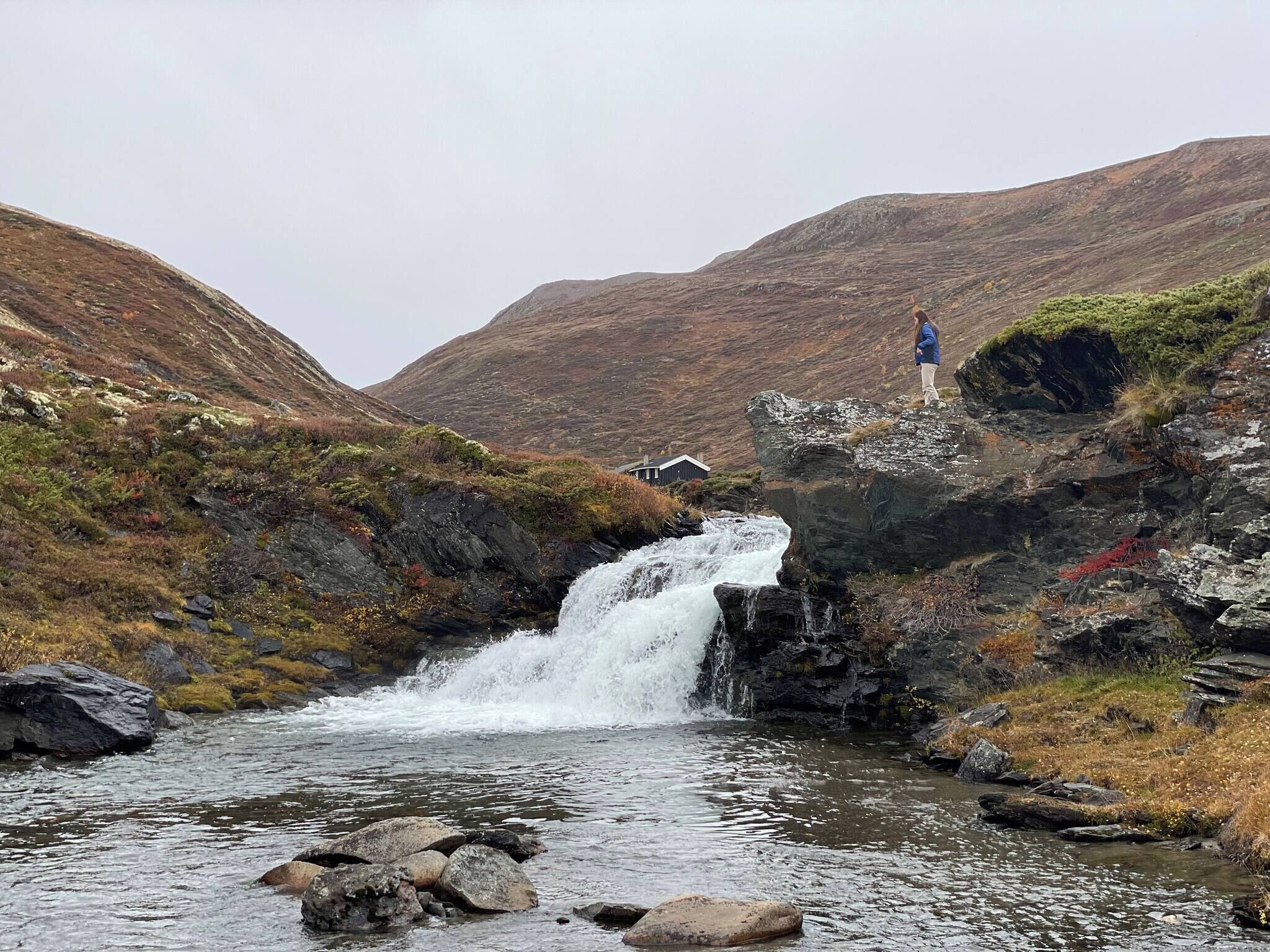 A waterfall in a mountainous area with a person standing on a rock.