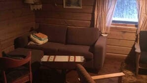 Een gezellig cabin interieur met een bruine bank, een houten stoel en een kleine tafel. Er is een raam met gordijnen zichtbaar, en een lamp hangt aan de muur.