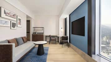 A modern living room with a sofa, a dining table, and chairs, a TV mounted on the wall, and a large window with a city view.