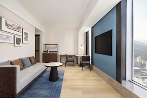 A modern living room with a sofa, a dining table, and chairs, a TV mounted on the wall, and a large window with a city view.