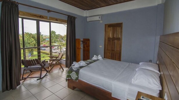 A bedroom with a large bed, a wooden ceiling, a window with curtains, a chair, and a view of greenery outside. - La Fortuna Downtown Boutique Hotel (La Fortuna)