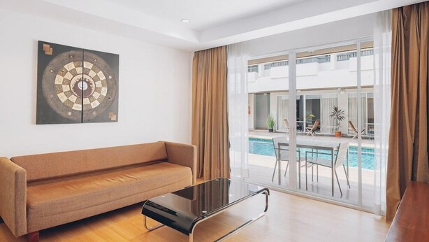 A modern living room with a sofa, a glass coffee table, and a large window with a view of a pool and outdoor seating. - Chateau Dale Boutique Residence (Pattaya)