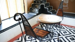 A dining area with a round table and chairs with wooden slats and metal frames, a patterned rug, and a window with blinds. - PRABANCH GARDENS - A CONSCIOUS STAY (Chengalpattu)