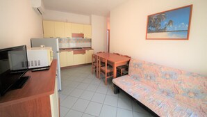 A compact kitchen with a refrigerator, microwave, and stove, a dining table with chairs, and a sofa with a patterned cushion. - Apartment in Rosolina for six guests (Rosolina Mare RO)