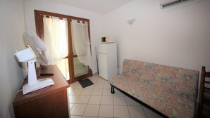 A small room with a wooden desk, a white fan, a microwave, a sofa, and a door with curtains. - Apartment in Rosolina for six guests (Rosolina Mare RO)
