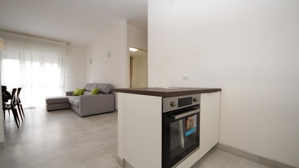 A modern kitchen with a built-in oven and a dining area with a grey sofa and green cushions. - Stylish Stay for 4 near Porto Caleri (Rosolina Mare RO)