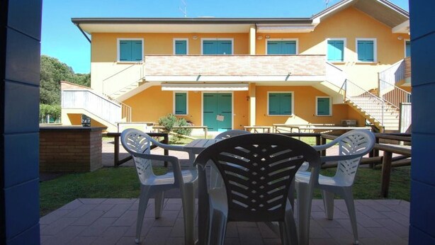 A two-story building with a yellow facade, a green door, and multiple windows. There is a patio area with outdoor furniture. - Family stay in Rosolina for six guests (Rosolina Mare RO)