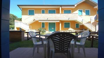 Un bâtiment à deux étages, doté d’une façade jaune, d’une porte verte et de nombreuses fenêtres. On y trouve une terrasse avec du mobilier d’extérieur.