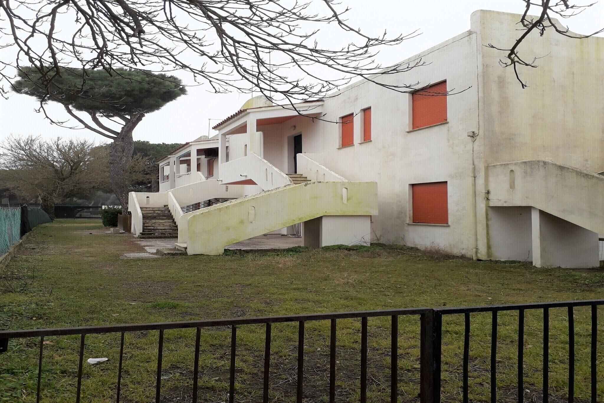 A building with a staircase leading to a covered entrance, surrounded by a grassy area and a tree.