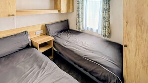 Ein kleines Schlafzimmer in einem Wohnmobil mit zwei Betten, einem kleinen Nachttisch aus Holz und einem Fenster mit gemusterten Vorhängen.