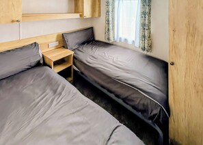 A compact RV bedroom with two beds, a small wooden nightstand, and a window with patterned curtains. - 2 bedroom accommodation in Haverigg, Millom (Haverigg, Millom)