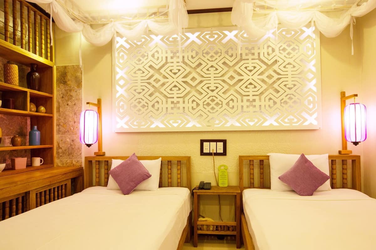A hotel room with two beds, a decorative wall panel, bedside lamps, a nightstand with a phone and a bottle, and a shelf with various items.