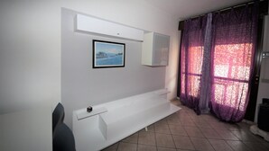 A room with a white bench, a framed picture on the wall, and a window with purple curtains. - Holiday Apartment in Rosolina Mare (Rosolina Mare)