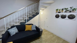 Un salon avec un canapé noir, un escalier et des œuvres d’art végétales accrochées au mur.