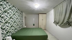 Ein Schlafzimmer mit grüner Bettdecke, einem weißen Nachttisch, einem Kleiderschrank und einer Wand mit einem Blattmuster.