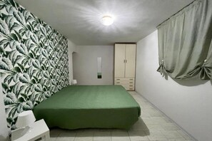 Een slaapkamer met een groen beddeksel, een witte nachtkastje, een kledingkast en een muur met een bladpatroon.