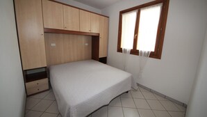 A bedroom with a bed, wooden wardrobe, and a window with curtains. - Apartment for Six in Rosolina (Rosolina Mare RO)