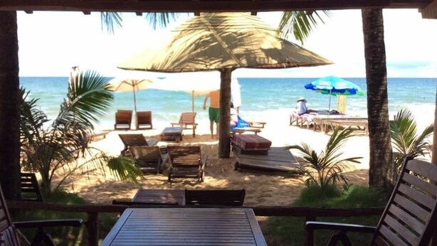 Blick auf einen Strand mit einer Holzterrasse, Liegestühlen, einem Strohdachschirm und einem Holztisch.
