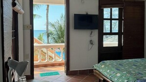 Ein Zimmer mit einem Bett, einem an der Wand befestigten Fernseher, einem Balkon mit Blick auf Palmen und einem Essbereich mit Stühlen und Tisch.