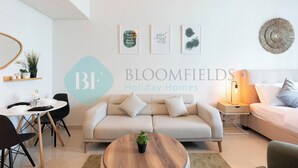 A modern living room with a sofa, a coffee table, and a dining area with chairs and a table. There are framed artworks on the wall and a logo for 'Bloomfields Holiday Homes'. - Bloomfields Spectacular STU Julphar (Abu Dhabi)