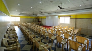 Una sala de conferències amb files de cadires de plàstic, un escenari amb catifa vermella i ventiladors de sostre.