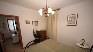 Ein Schlafzimmer mit einem Bett, einem Schreibtisch, einem Stuhl, einem kleinen Tisch mit Lampe, einem Spiegel und einer Tür zu einem anderen Raum.