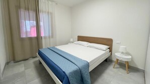 A single bed with a wooden headboard, a bedside table with a lamp, and a window with sheer curtains. - Cozy Apartment in Rosolina for 4 (Rosolina Mare RO)