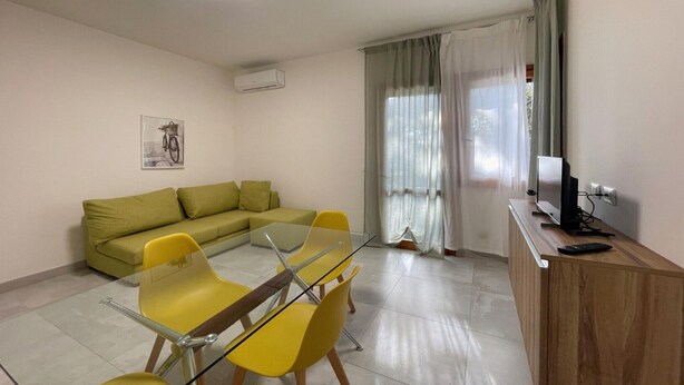 A living room with a sofa, a glass table, yellow chairs, a TV, and a wooden cabinet. - Cozy Apartment in Rosolina for 4 (Rosolina Mare RO)
