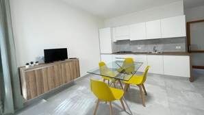 A modern kitchen with white cabinets, a glass dining table, and yellow chairs. - Cozy Apartment in Rosolina for 4 (Rosolina Mare RO)