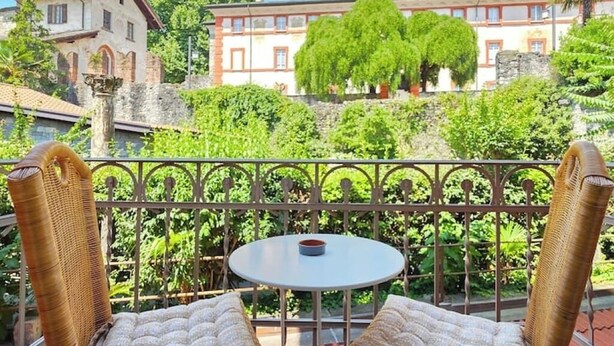 Un balcón con sillas de mimbre y una pequeña mesa, con vistas a un jardín con árboles y un edificio al fondo.