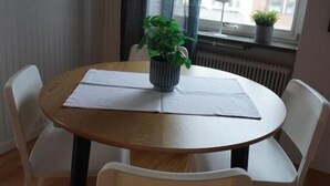 Ett runt träbord med två vita stolar, en krukväxt och ett fönster med utsikt över byggnader.