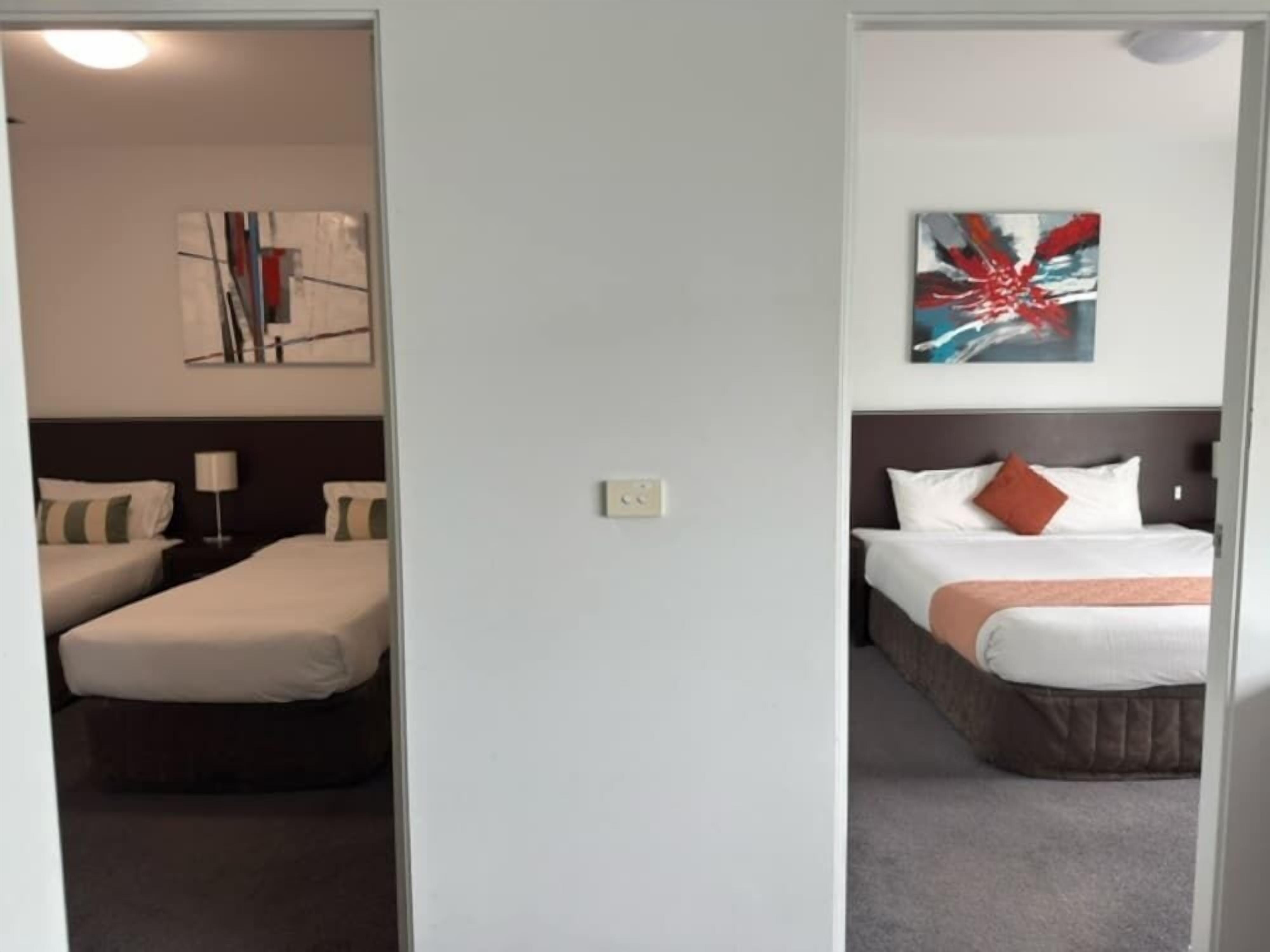 Une chambre d’hôtel avec deux lits, une table de chevet, une lampe et des œuvres d’art abstraites accrochées au mur.