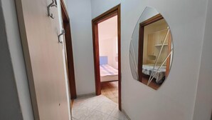 Un corridoio stretto con una porta che dà su una camera da letto. La stanza ha un letto con copriletto a scacchi e un piccolo specchio rotondo appeso al muro.