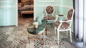 A room with a patterned floor, a green table, two patterned chairs, a bench, and a brick wall. - Acronafplia Pension B&D (Nafplio)