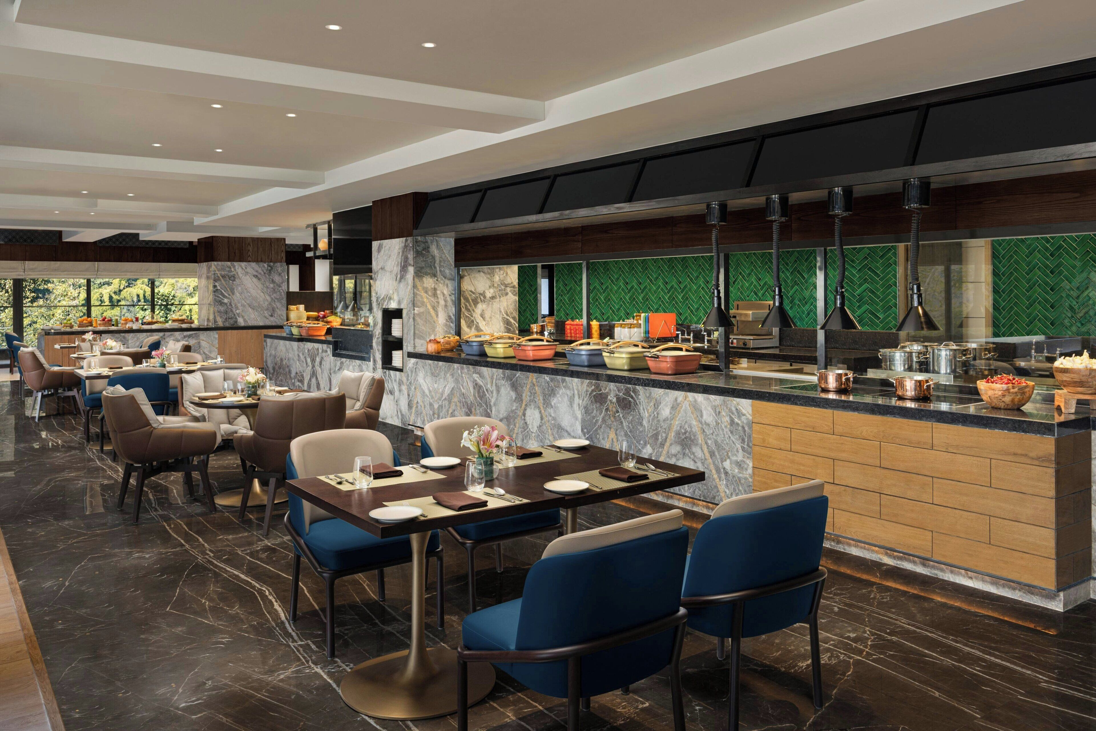 Un restaurante moderno con pisos de mármol, sillas tapizadas en azul y una zona de cocina con un revestimiento verde tipo espiga.