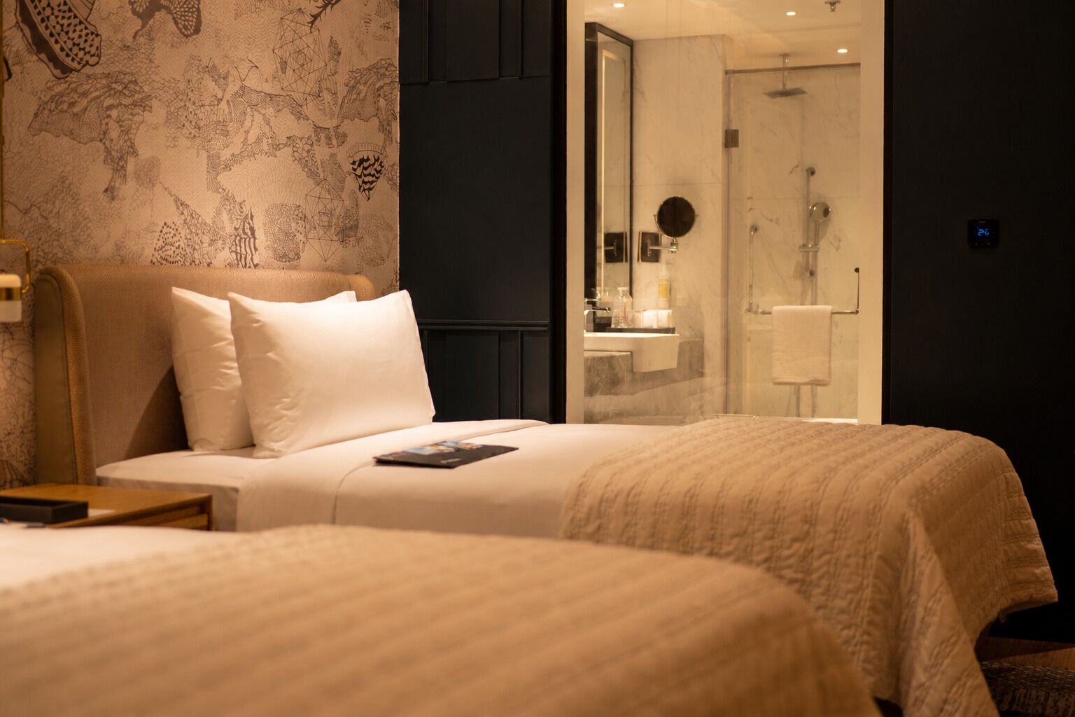 A hotel room with two beds, a bathroom with a glass shower, and a wall with a geometric pattern.