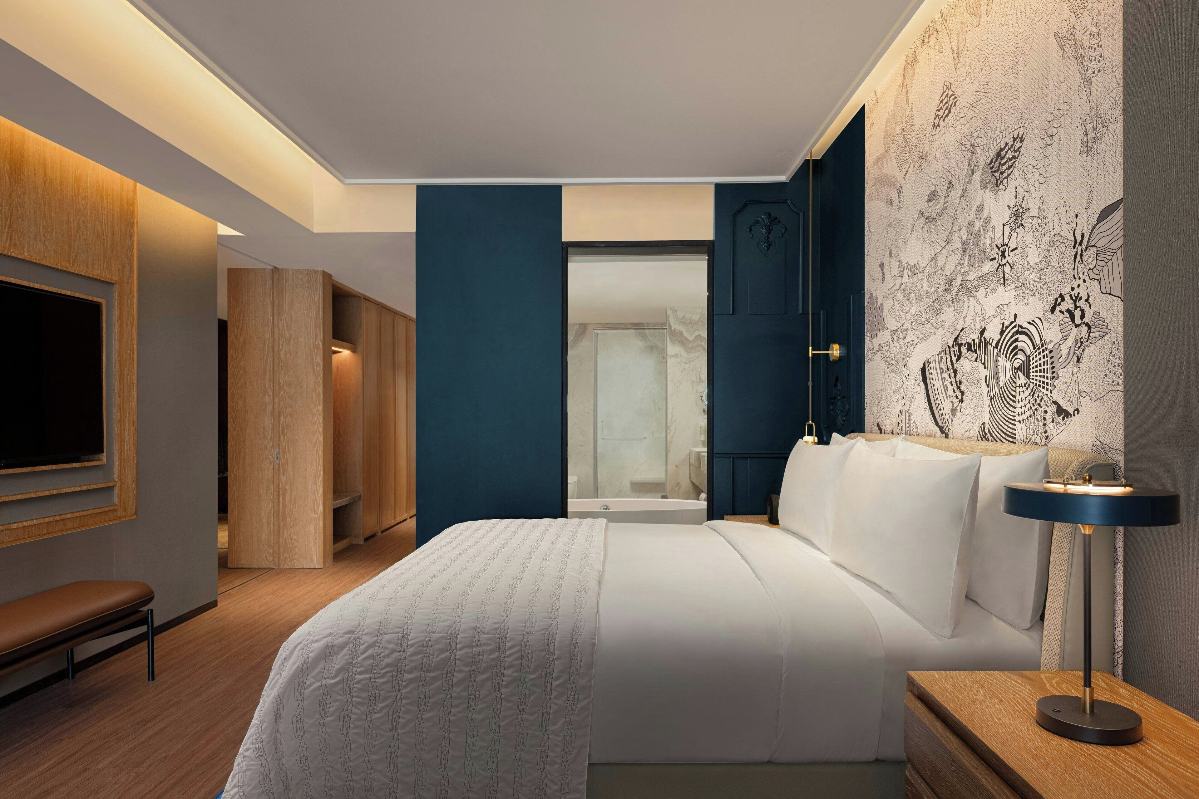 A modern hotel room with a large bed, a bedside table with a lamp, a wardrobe, and a bathroom visible through an open door.